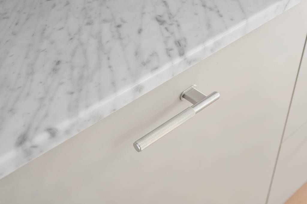 Närbild på ett handtag i rostfritt stål monterat på en ljusgrå kökslucka med bänkskiva i marmor.