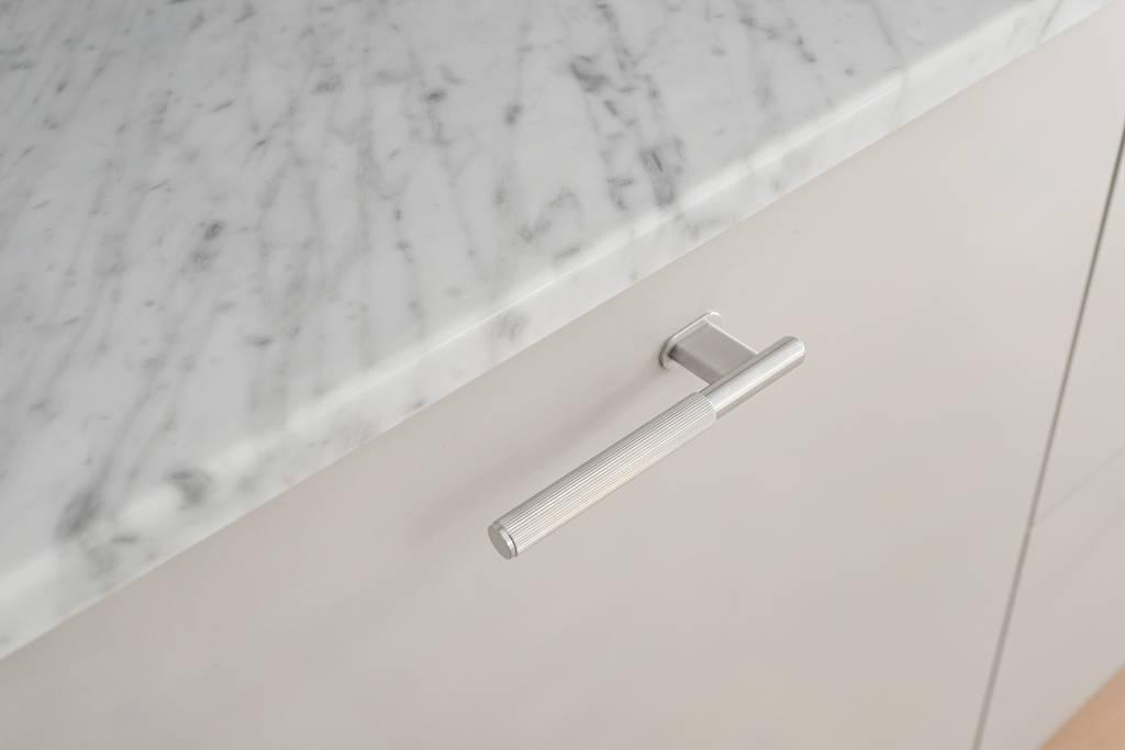 Närbild på ett handtag i rostfritt stål monterat på en ljusgrå kökslucka med bänkskiva i marmor.