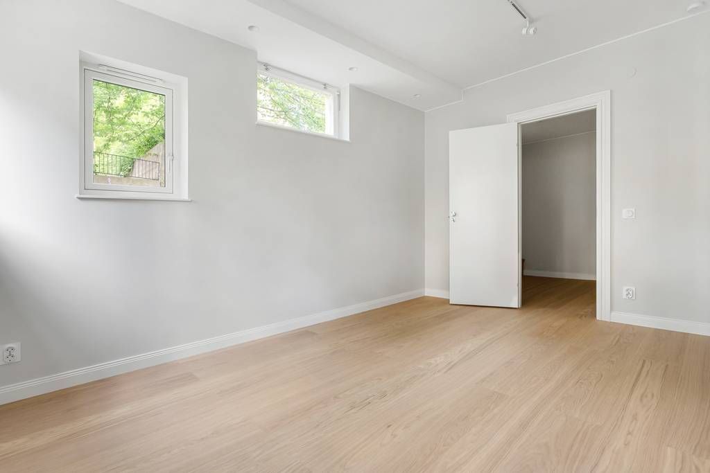 Allrum med fönster som vetter mot grönska, vitmålade väggar och ljust parkettgolv.